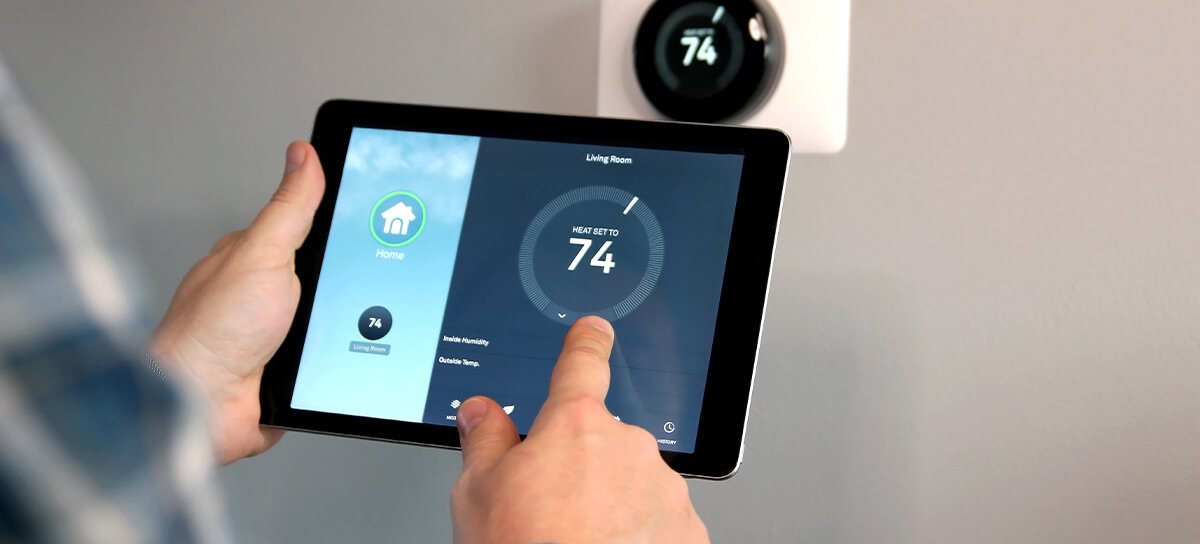 Man holding smart thermostat tablet with smart thermostat in the background on wall.